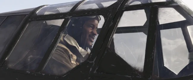 Film still of a smiling WWII pilot seen through the framed glass canopy of a dark fighter aircraft