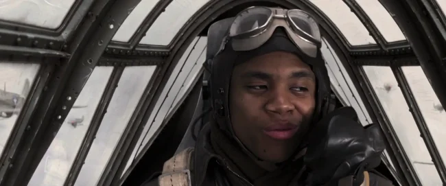 Film still of a young WWII fighter pilot in leather flight helmet and goggles smiling inside a glazed cockpit canopy