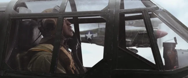 Film still shot through a weathered cockpit canopy of a WWII pilot in profile, a star-marked fighter flying alongside