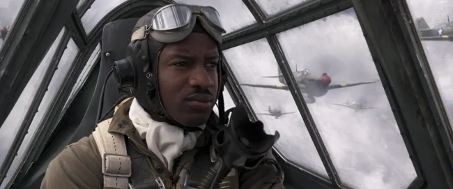 Film still of a Black fighter pilot in leather helmet and goggles pointing from a cockpit, warplanes visible outside