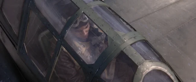 Overhead film still of a pilot in flight helmet and goggles seated under the closed glass canopy of a fighter plane