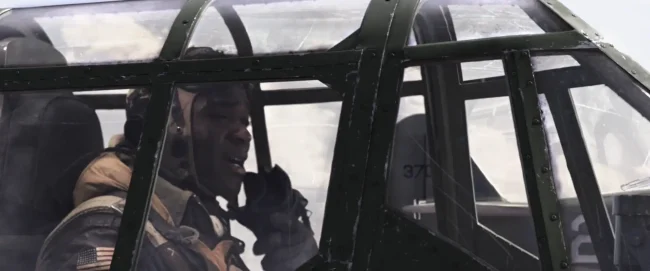 Film still of a WWII pilot with US flag patch speaking into an oxygen mask inside a framed cockpit canopy