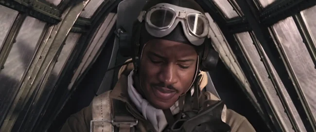 Film still of a WWII fighter pilot in leather helmet and goggles grimacing in a riveted cockpit canopy